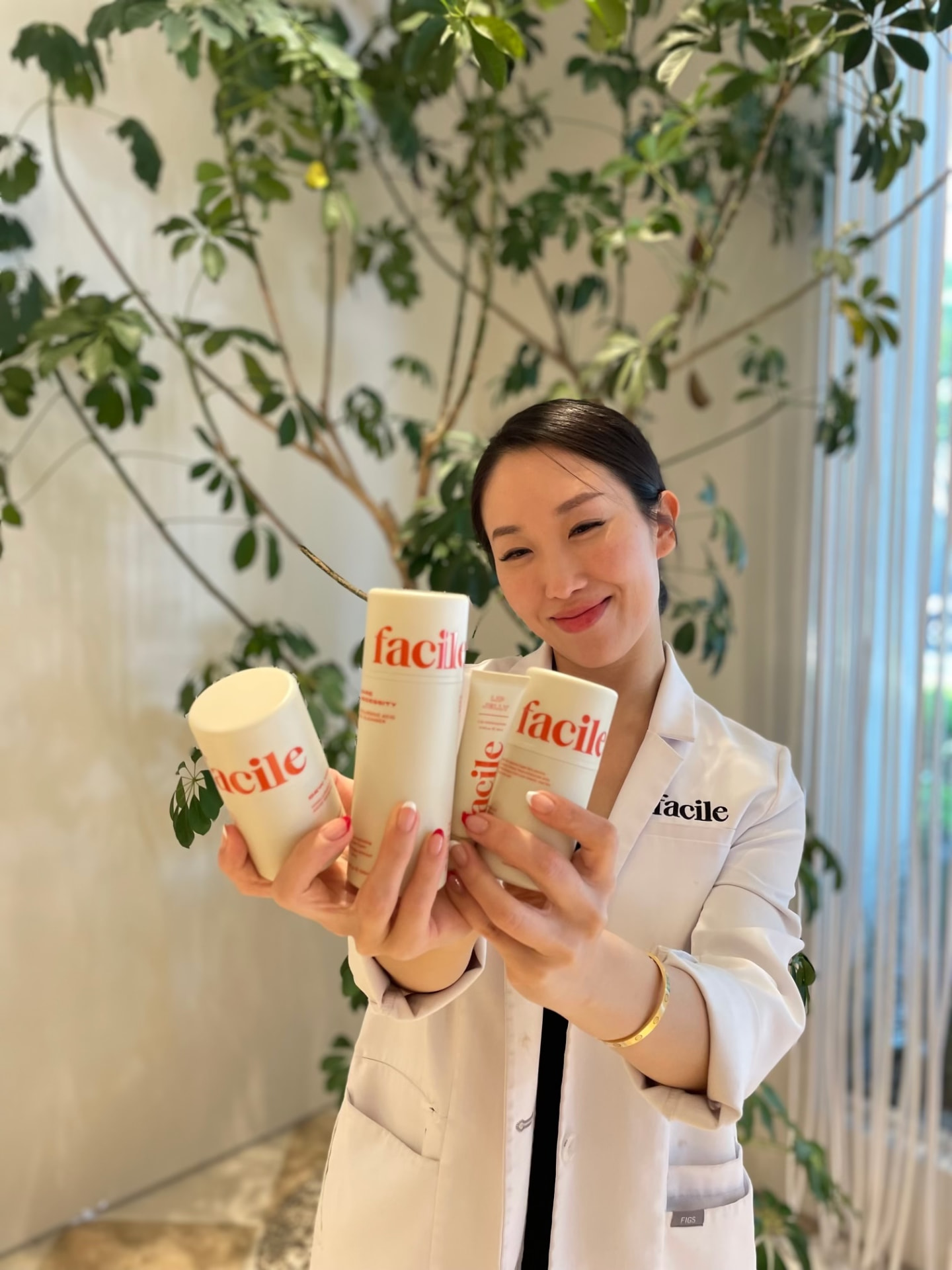 Facile esthetician smiling and holding four Facile products up to the camera in front of a plant