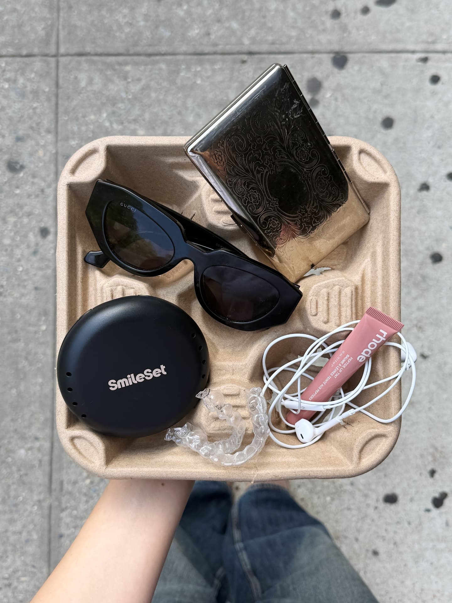 Flatlay tray with sunglasses, a vintage case, aligners, and lip tint on a city sidewalk