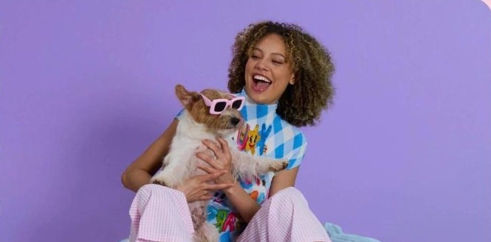Woman laughing with a corgi in pink sunglasses against a lavender backdrop